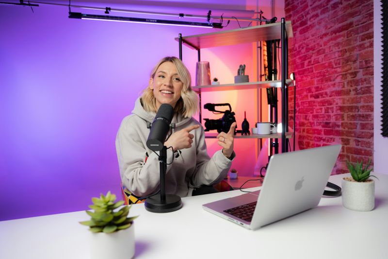 A person in hoodie smiling and pointing at desk setup with mic, laptop, and plants.