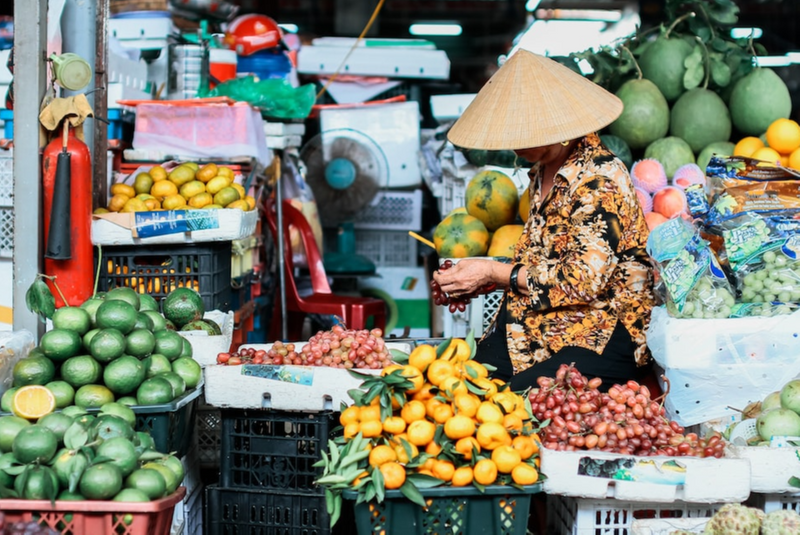 A woman in a pointed straw hat picking over tropical fruit at a traditional market.