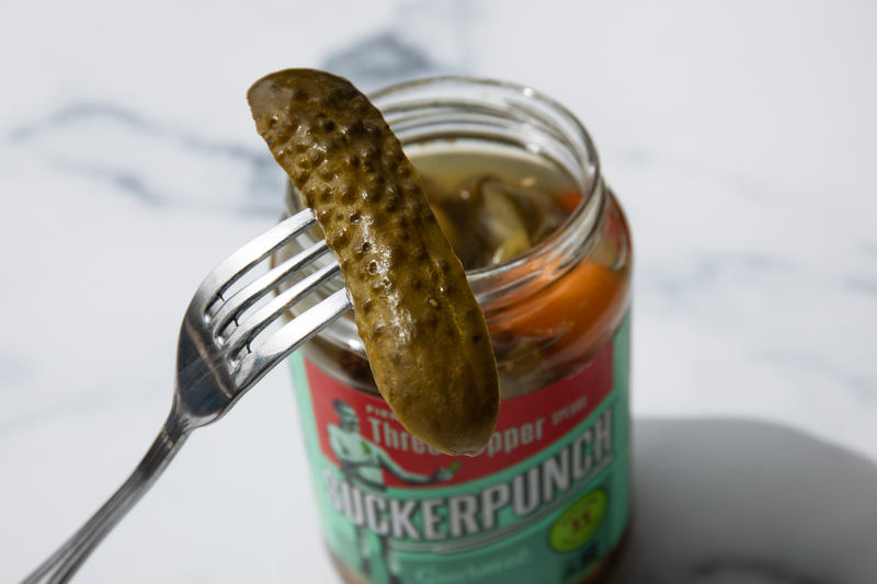 A fork with a pickle on it and a jar of pickles sitting on a white counter in the background.