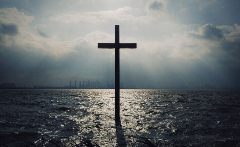 Large cross jutting out of the ocean with city skyline and clouds in the background.