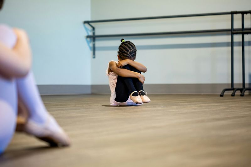 A girl curled up in the corner of a ballet class, curled into a ball.