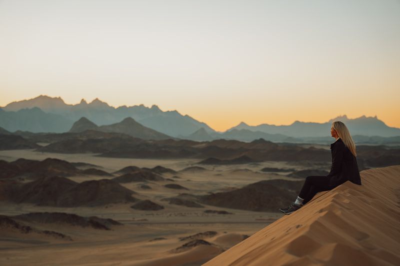 A woman sitting on a sand dune, looking out at a rocky desert landscape.