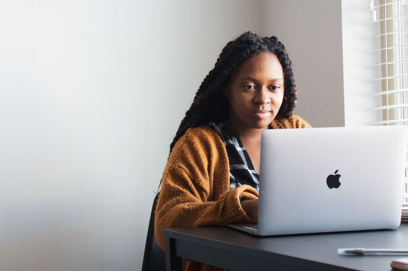 Young Black woman with long hair and orange sweater working on laptop.