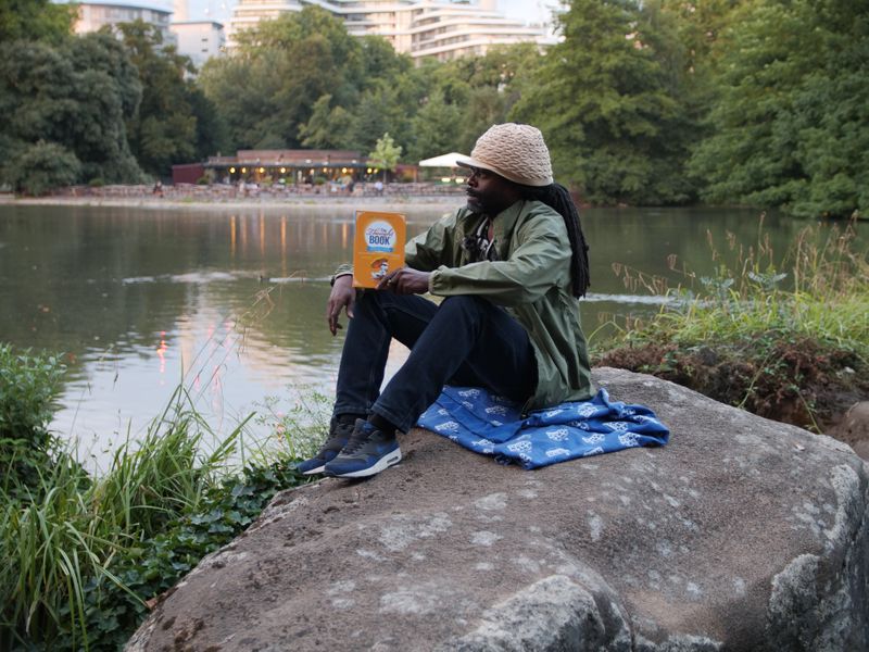 Man reading a book on a rock by a lake, with a town in the far background