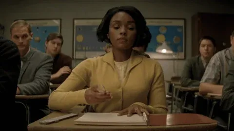 A girl with a serious expression sits at a classroom desk, focused on taking notes.