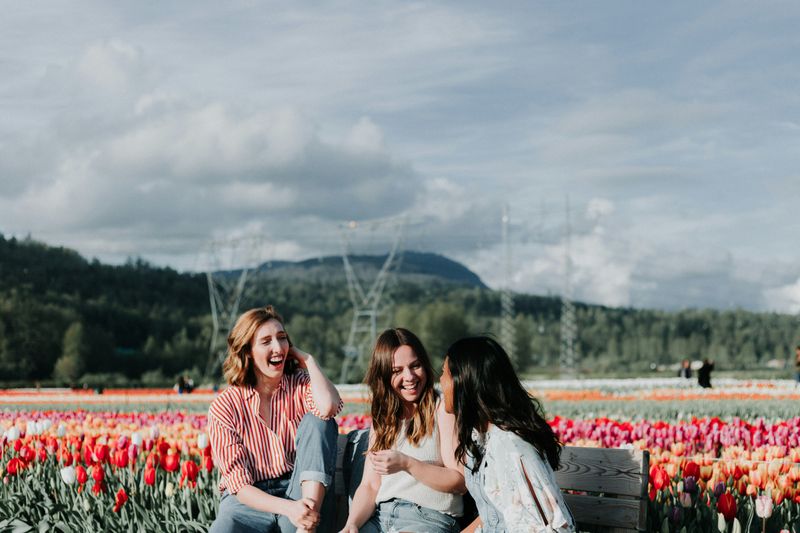 Three women smiling while sitting on a bench in front of a field of red, pink, and orange tulips. 