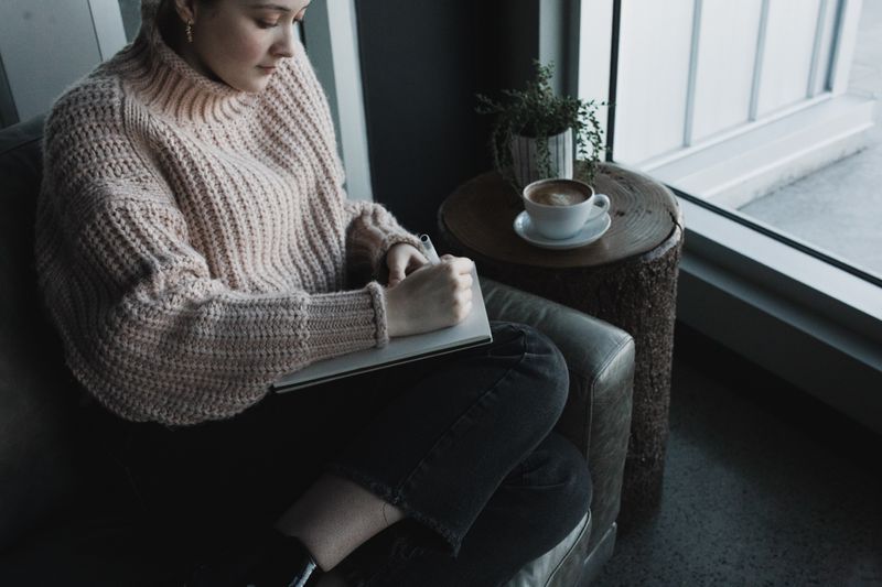 A woman in a sweater sits on a leather chair near a window journaling. A cup of coffee rests on a small table beside her.