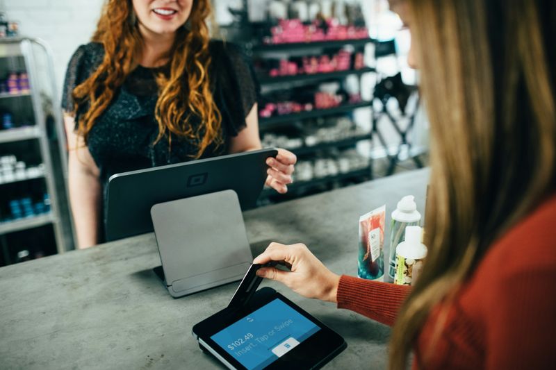 Two women in a beauty store. One stands behind the counter and the other taps their card to make a purchase.