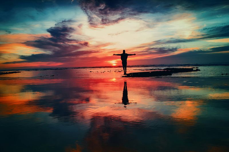 A person standing on a rock in the middle of water looking at a beautiful, colorful sunset.