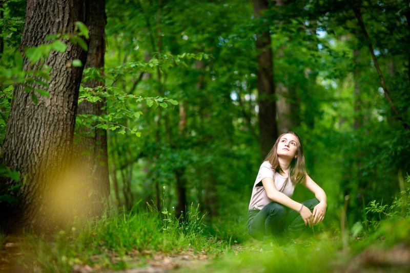 A woman in a forest. She sits in a crouched position and looks up towards the sky.