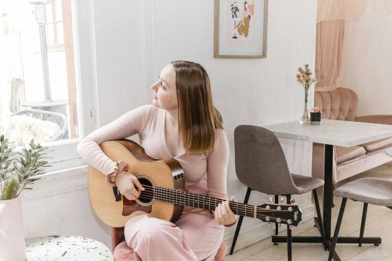 A women playing the acoustic guitar at home, looking out a window.