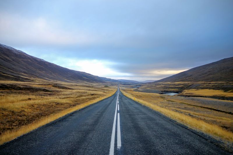 An open stretch of road with no vehicles with mountains and grassland in the surrounding area.