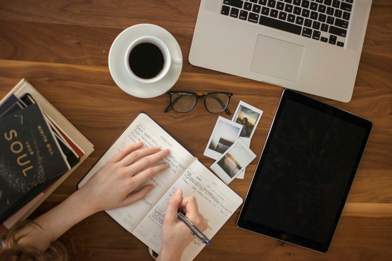 A person practicing for the IELTS Writing test. They write in a notebook on their desk in front of a laptop.