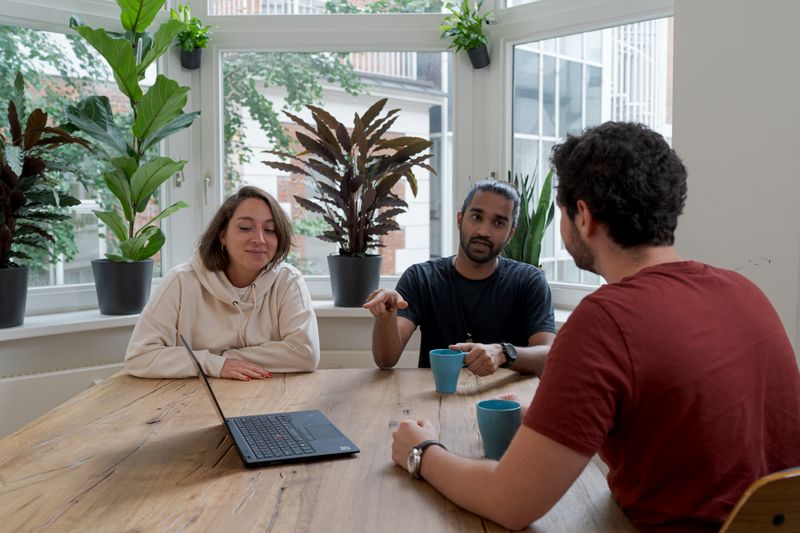 Two men and a woman sitting around a table talking and looking at a laptop. In the background are several potted plants.