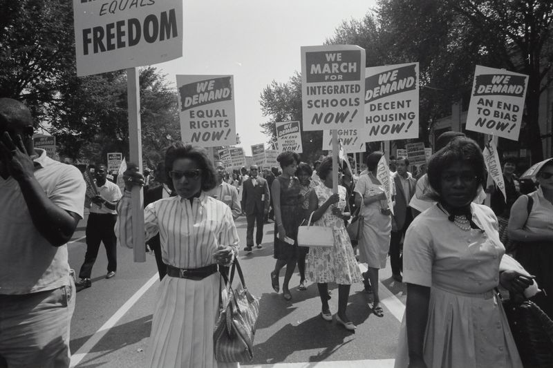 A Civil Rights march in the 1960's. People are proceeding down a street, holding signs demanding equality..