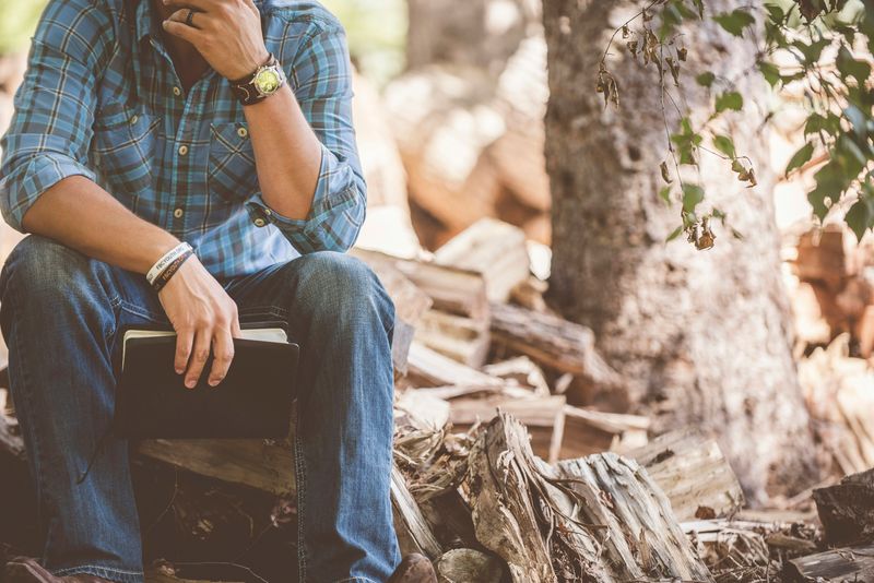 A man holding a notebook sitting on a tree stump in a forest. 