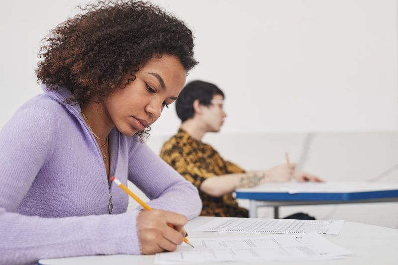 A young woman sitting at a desk in an exam room, writing on her test paper with a pencil.