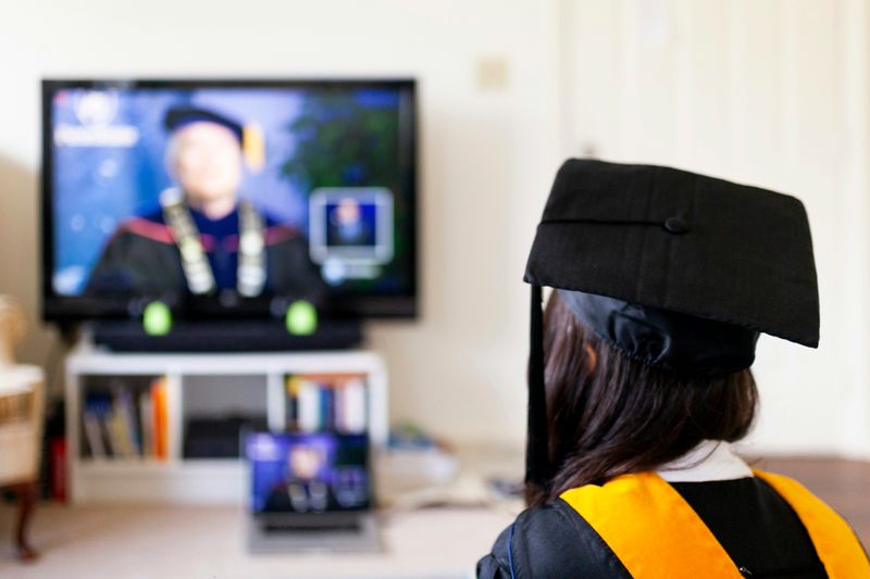 A graduate in their cap and gown looks at their professor (also in graduation clothes) on a TV screen.