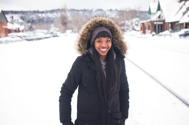  woman wearing a black snow jacket, gray scarf, and purple hat in front of a white, snowy background