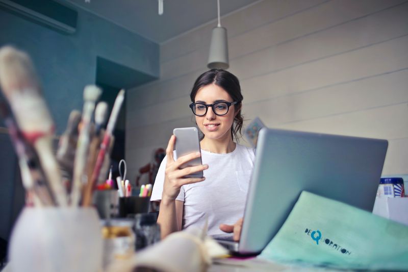 A woman at a cluttered desk with a laptop checks her smart phone.