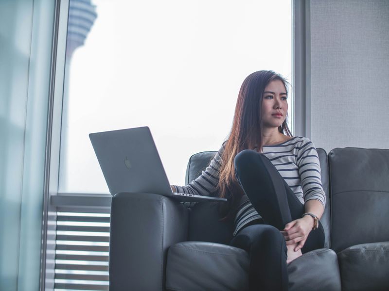 A woman sitting on a couch with a laptop. She looks away in thought.