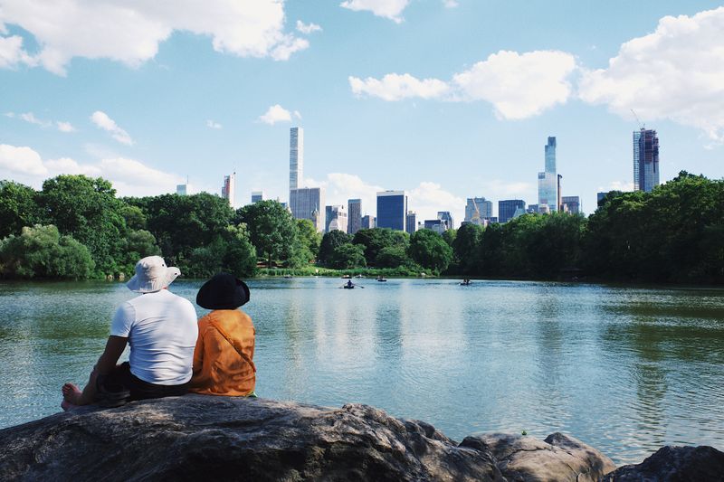 Couple sit facing the lake in Central Park, New York City, with the city skyline above the distant tree line.