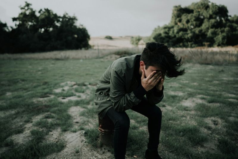 A young man sitting alone with his head in his hands. His fingers are covering his eyes. 