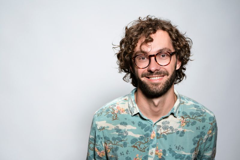 A man in mid 30's with curly brown hair, beard, and brown glasses. He is wearing a blue button shirt with trees and clouds.
