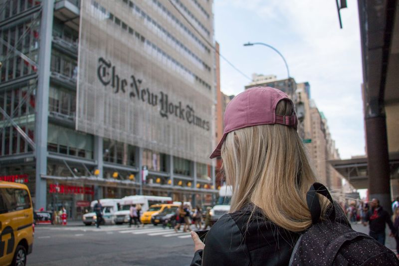 Image: Person in baseball cap looks at mobile phone with The New York Times headquarters in the background.