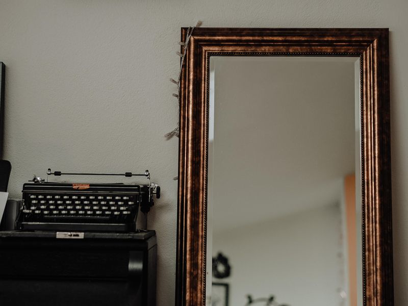 A mirror next to a typewriter and shelf reflects back objects on the other side of a room.
