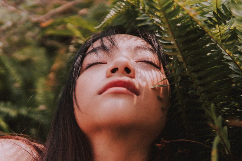 A woman under a fern closes her eyes.