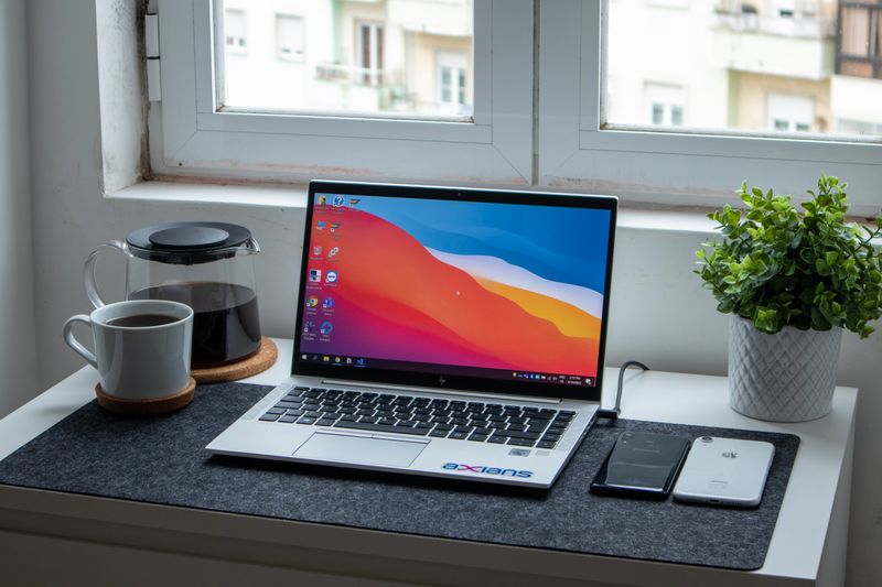 An open laptop sits on a desk in front of a sunny window. alongside a pot of coffee and a small plant.