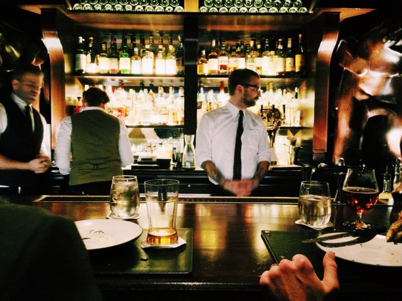 A bartender stands at a bar with a beer and an empty plate. There is alcohol behind him on the wall.