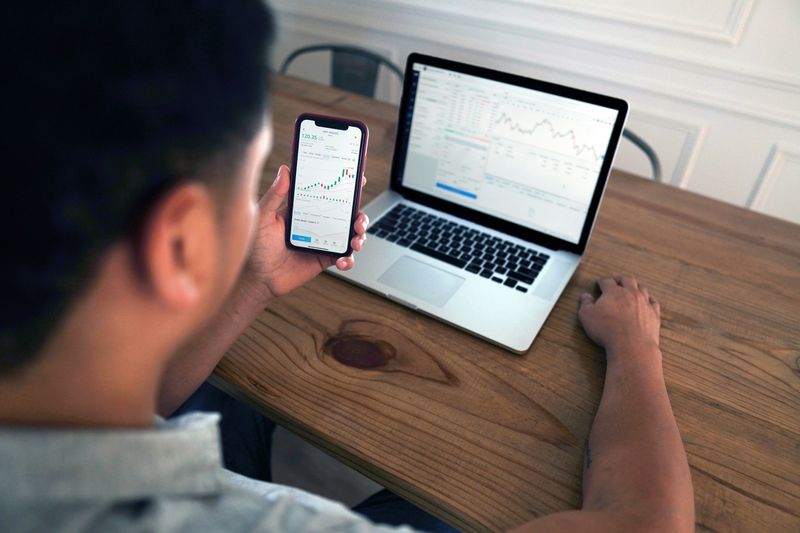 A person looking at their finance portfolio earnings on a phone app while looking at market trends on a laptop.
