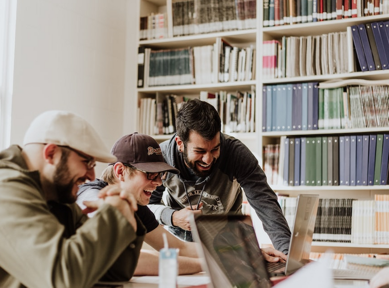 Three students are seen chuckling noticing something on their laptops. There is a large bookshelf to their left.