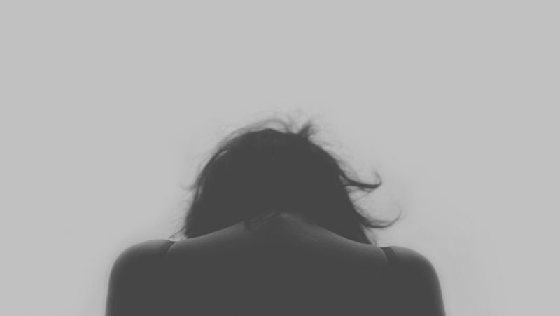The back of a woman with her head bowed low against a plain grey background.