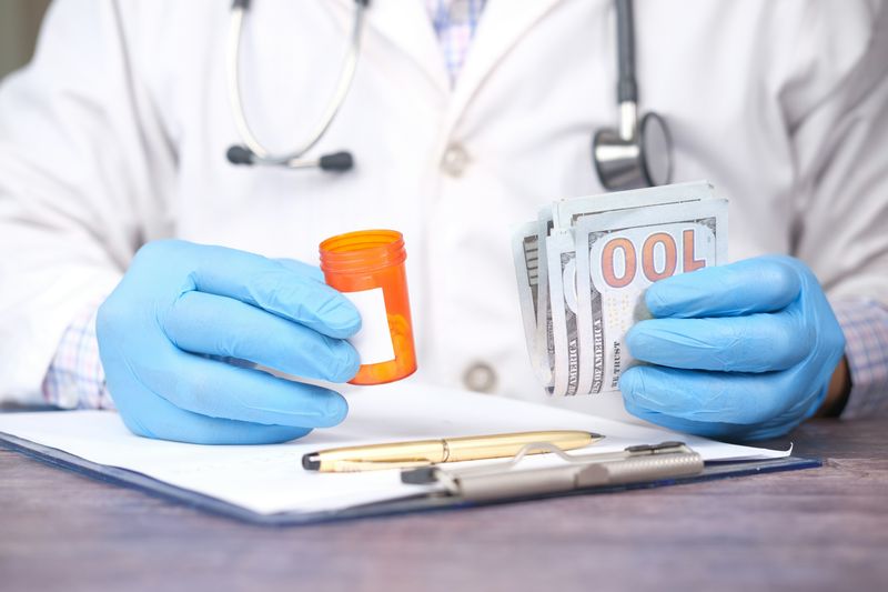 A doctor wearing gloves holding a prescription bottle in one hand and a stack of cash in the other, symbolizing medical costs