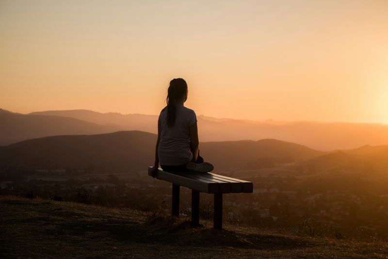 A woman sitting on a bench overlooking a mountain side at sunrise.