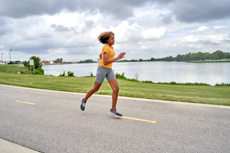 a woman wearing an orange shirt and gray shorts, jogging in a clear street with a grass and lake background
