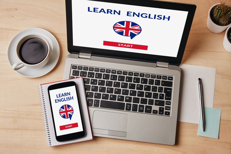 Laptop and phone on table with screens imprinted &apos;Learn English&apos; alongside notepad, pen, and coffee cup.