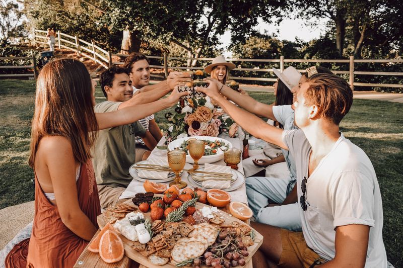 A group of men and women are sitting around a feast of food on grass. They are raising their glasses for a toast.