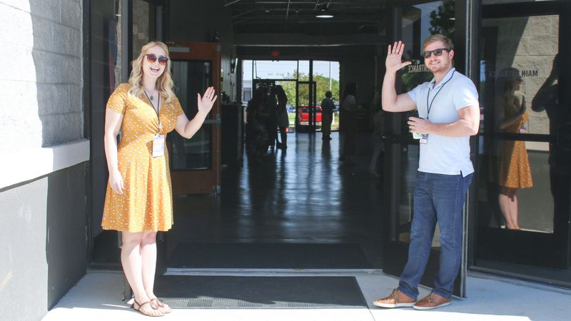 Two teachers waving while standing in front of open school doors.