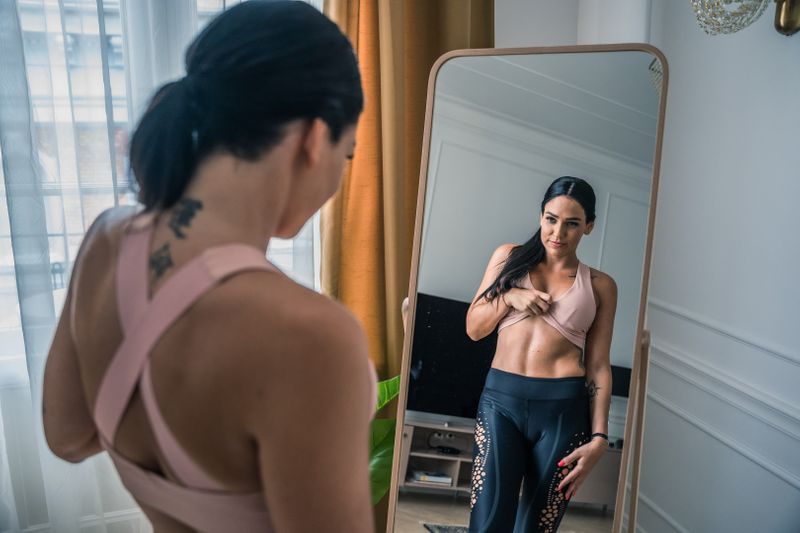 A woman is looking at herself in the mirror in her room, wearing a sports bra and sports leggings.