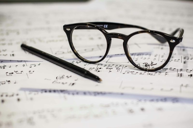 A pile of music sheets on a desk. A music education's student's glasses and a pen sit on the sheets.