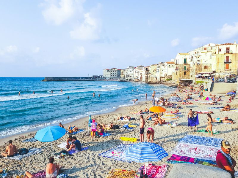 A beach scene with blue water filled with people swimming and others relaxing. Houses are displayed in the back.