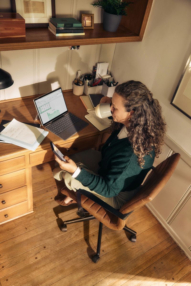 A woman is drinking from a mug in her office while sitting on a chair.