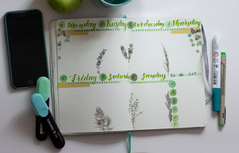 A phone and notebook calendar with light blue and green pens and markers surrounding it.