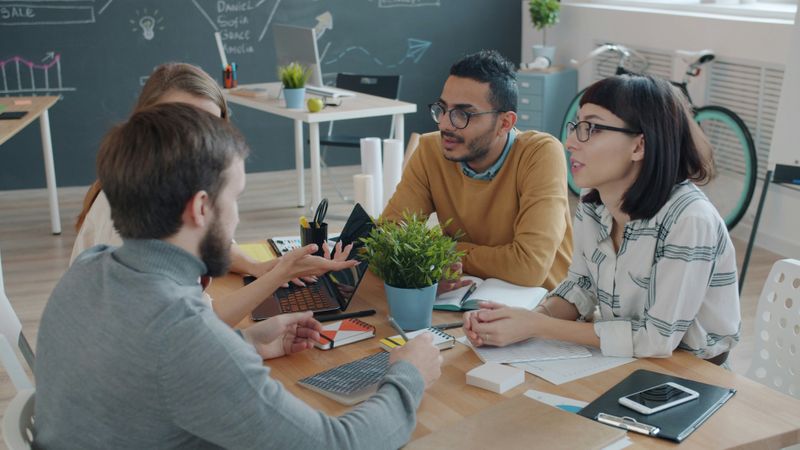 Four people sitting around a desk with a laptop, smartphone, and notebooks on the table. 