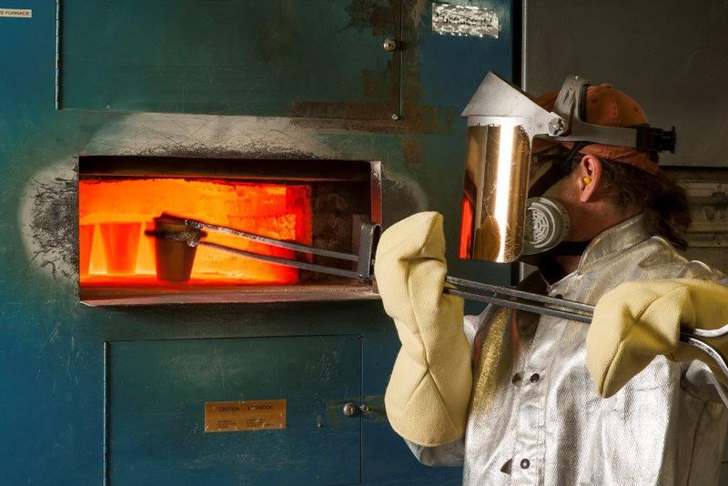 A metallurgist conducts a fire assay, putting ore into a furnace with large tongs.