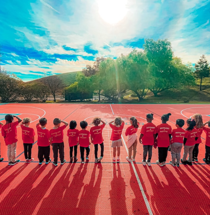 Elementary children, wearing red shirts and facing away from the camera, standing in line on an outside basketball court. 